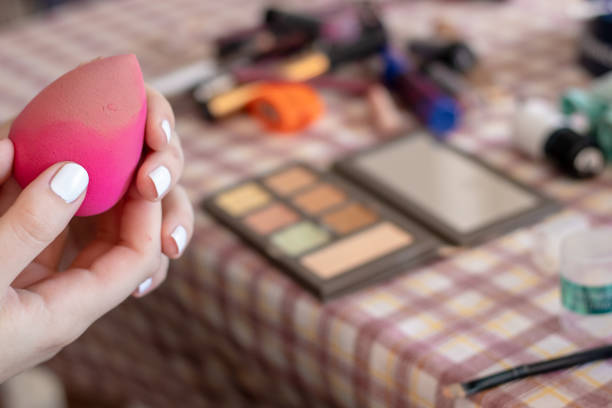 Female hands with white fingernails holding a pink make up sponge. Daily routine of applying make up, accessories and brushes in the background