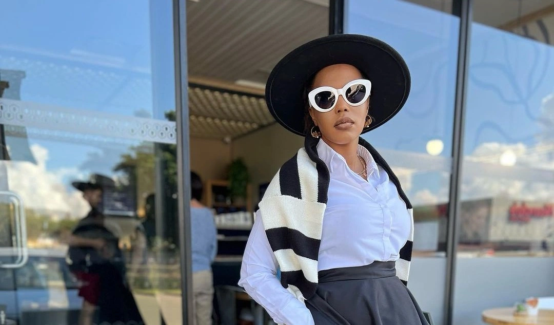 A fashion black woman in a white button-down shirt, midi skirt, fedora hat, white framed sunglasses, and high heels