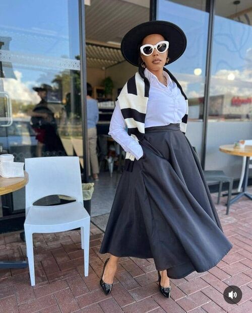 A fashion black woman in a white button-down shirt, midi skirt, fedora hat, white framed sunglasses, and high heels